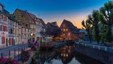 A charming European old town with colorful historic buildings, cobblestone streets, a riverside promenade, and a scenic castl
